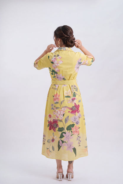 A woman viewed from behind wearing a yellow midi dress with a colorful floral print, standing against a plain light gray studio background.