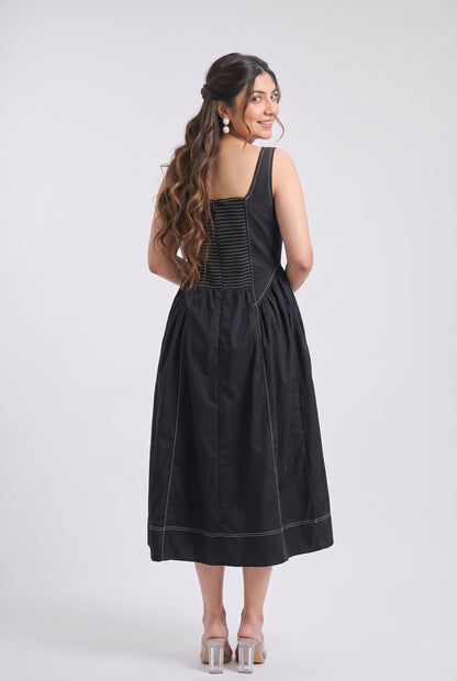 A woman modeling a black sleeveless midi dress from the back, featuring a smocked bodice and gathered skirt, set against a plain light gray studio background.