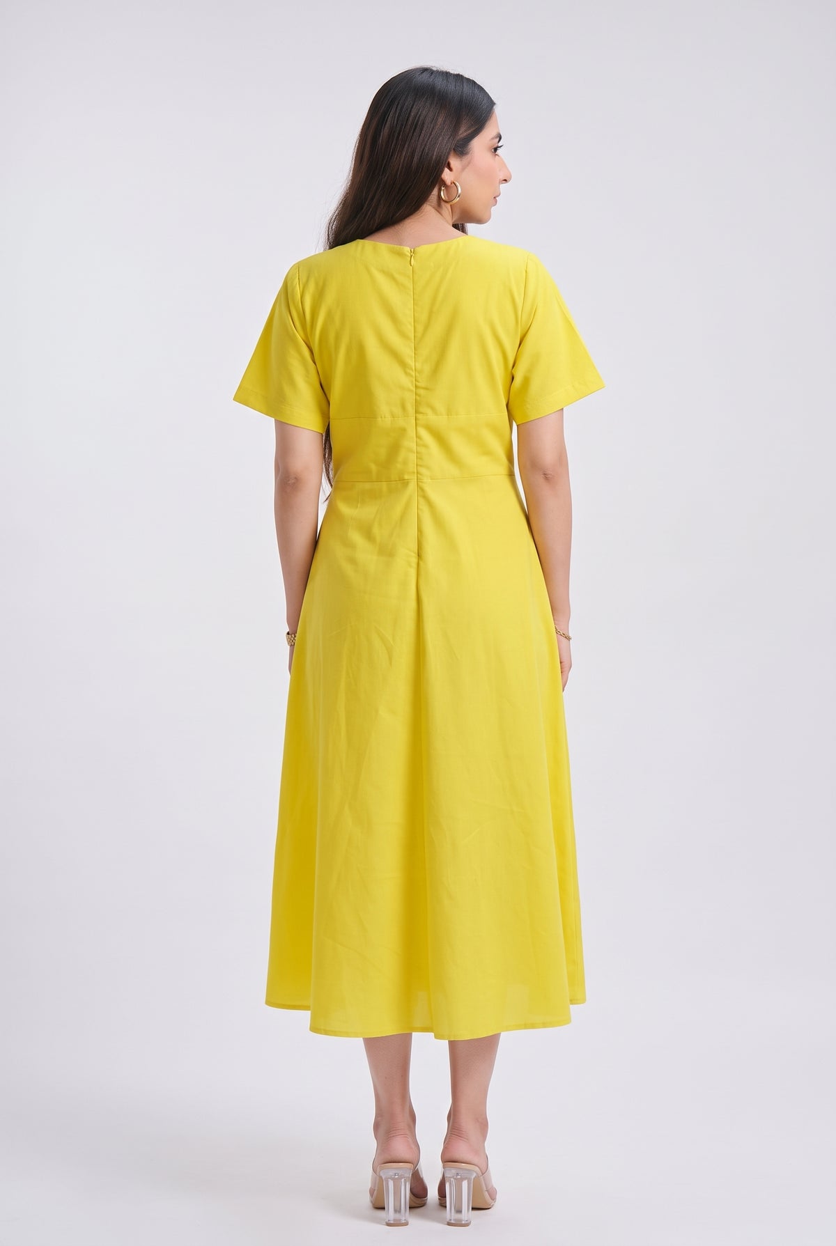 A woman modeling a bright yellow midi dress with short sleeves, a V-neckline, and a matching fabric belt with two gold-tone buttons, set against a plain light gray studio background.