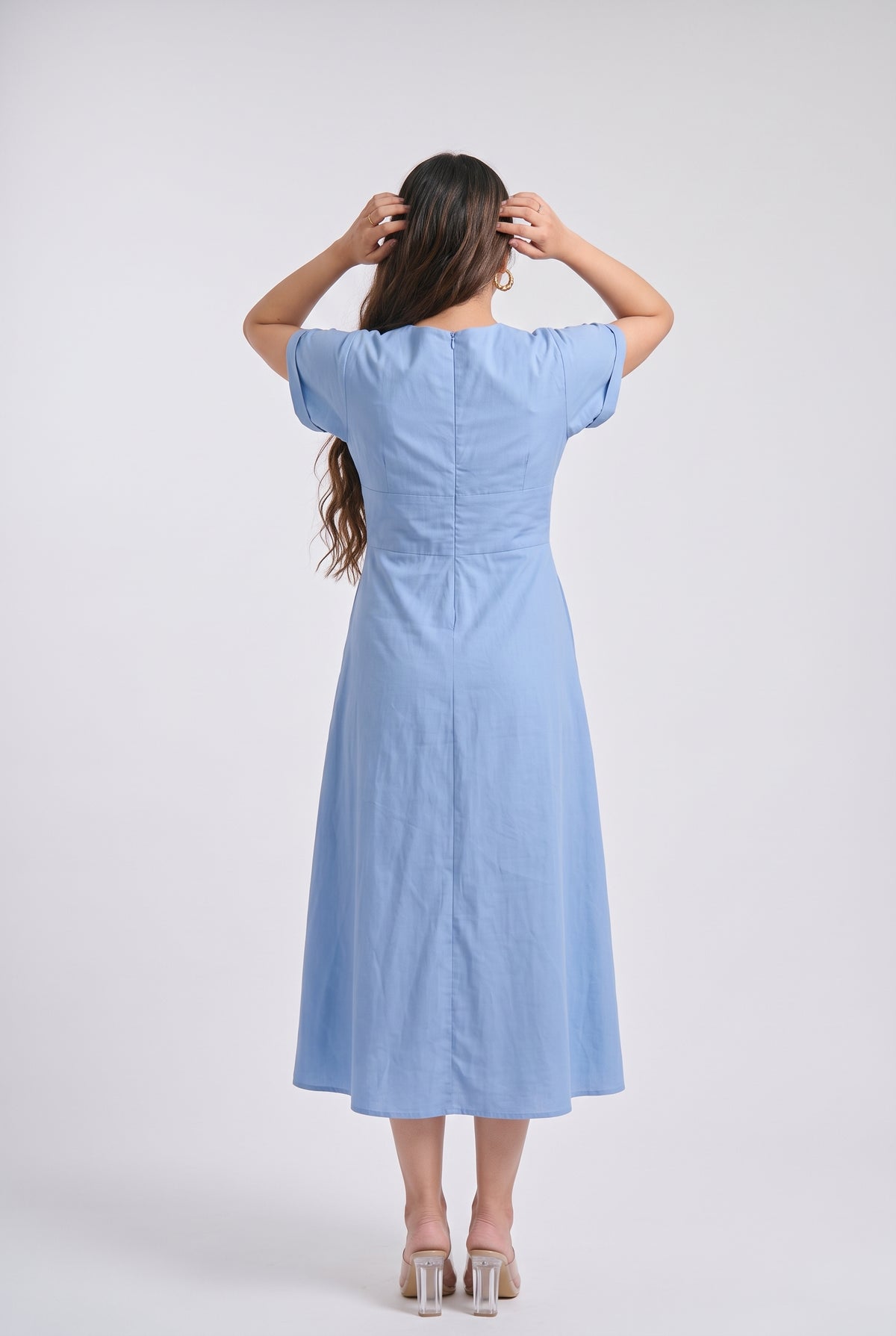 A woman modeling a bright Blue midi dress with short sleeves, a V-neckline, and a matching fabric belt with two gold-tone buttons, set against a plain light gray studio background.