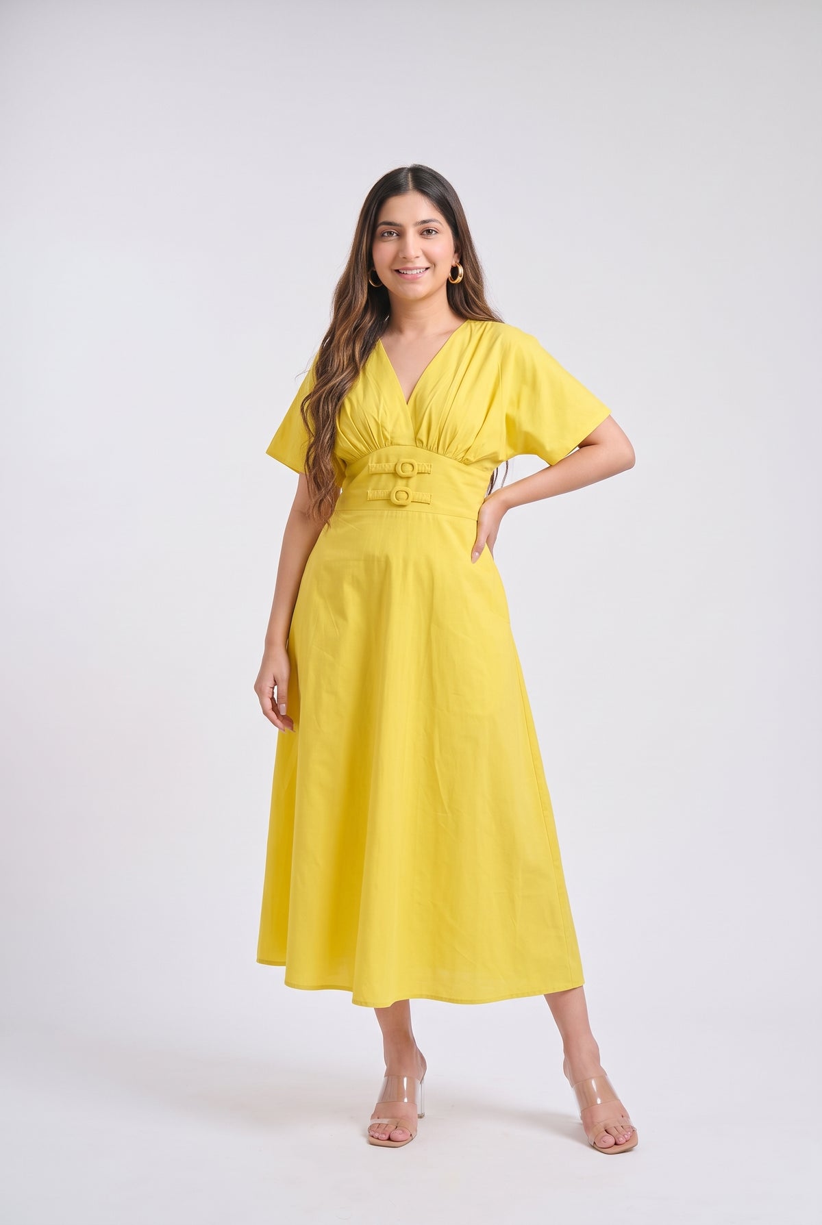 A woman modeling a bright yellow midi dress with short sleeves, a V-neckline, and a matching fabric belt with two gold-tone buttons, set against a plain light gray studio background.