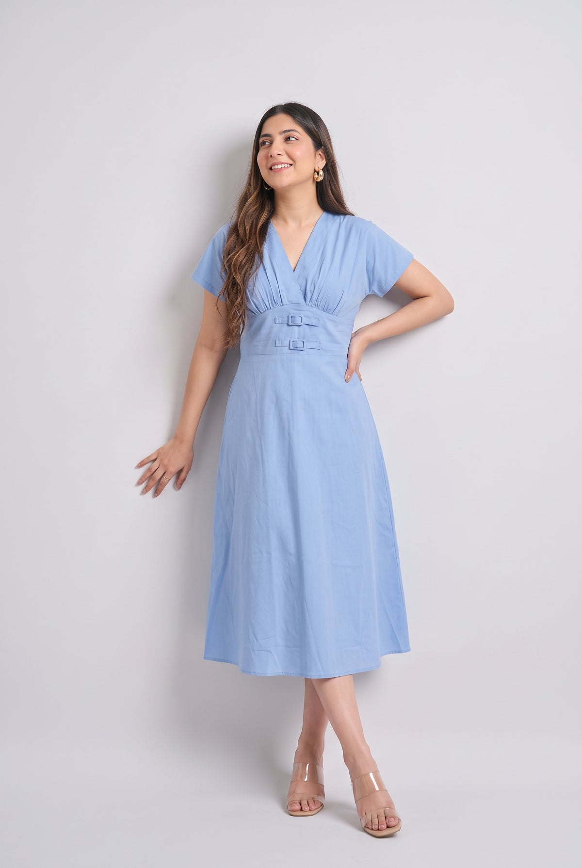 A woman modeling a light blue midi dress with short sleeves, a V-neckline, and a gathered waist detail, standing against a plain light gray studio background.