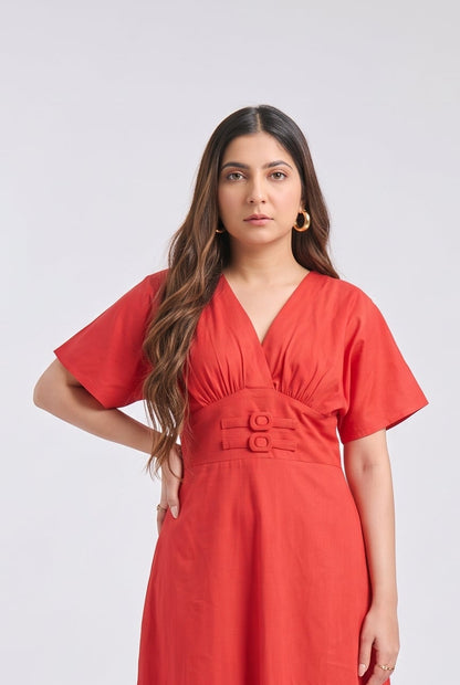 A woman modeling a bright Red midi dress with short sleeves, a V-neckline, and a matching fabric belt with two gold-tone buttons, set against a plain light gray studio background.