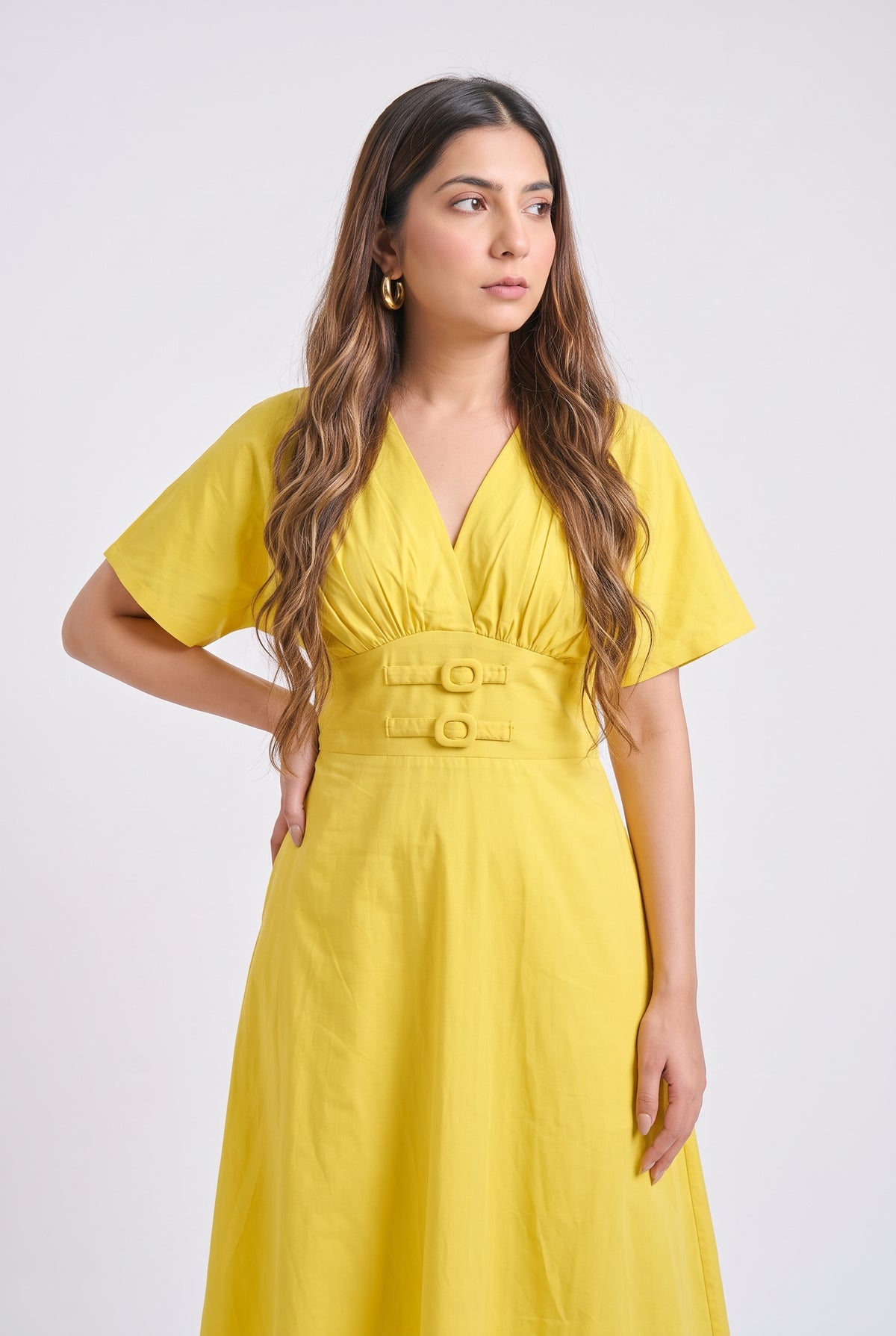 A woman modeling a bright yellow midi dress with short sleeves, a V-neckline, and a matching fabric belt with two gold-tone buttons, set against a plain light gray studio background.