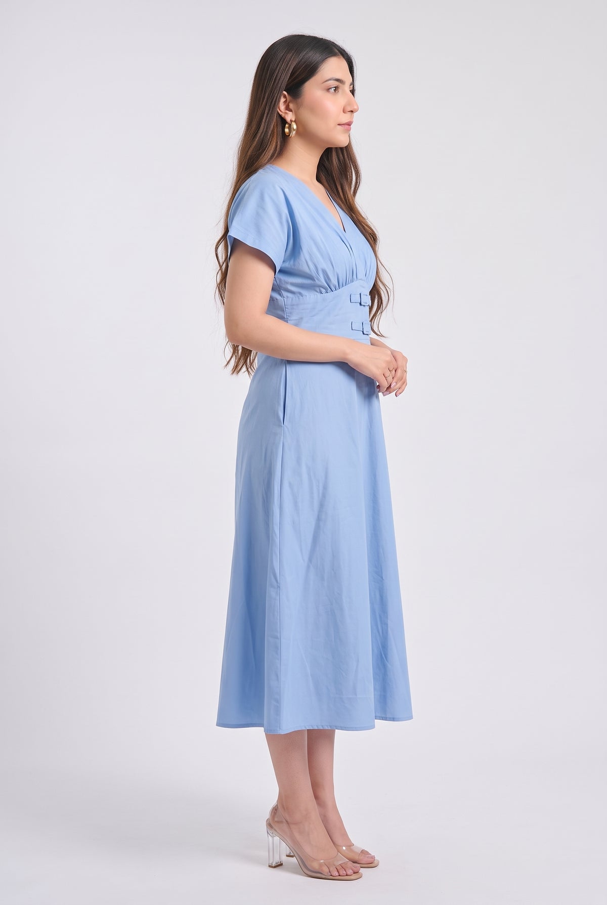 A woman modeling a light blue midi dress with short sleeves, a V-neckline, and a gathered waist, shown in profile against a plain light gray studio background.