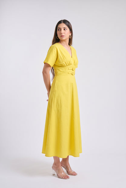 A woman modeling a bright yellow midi dress with short sleeves, a V-neckline, and a matching fabric belt with two gold-tone buttons, set against a plain light gray studio background.