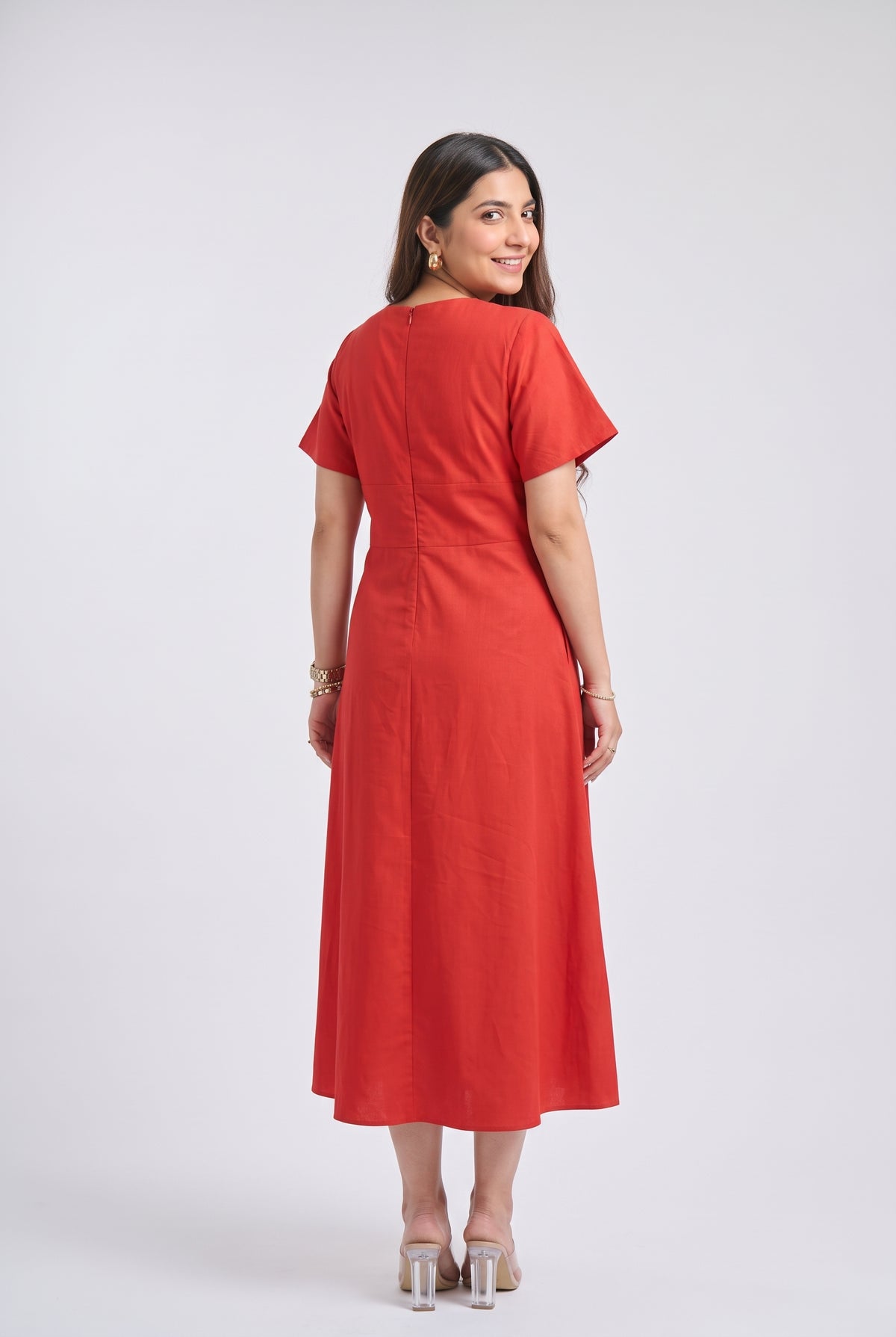 A woman modeling a bright Red midi dress with short sleeves, a V-neckline, and a matching fabric belt with two gold-tone buttons, set against a plain light gray studio background.