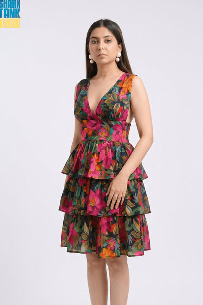 A woman modeling a sleeveless tiered dress with a vibrant floral print in pink, orange, and green, set against a plain light gray studio background. The 'SHARK TANK INDIA' logo is visible in the top left corner.