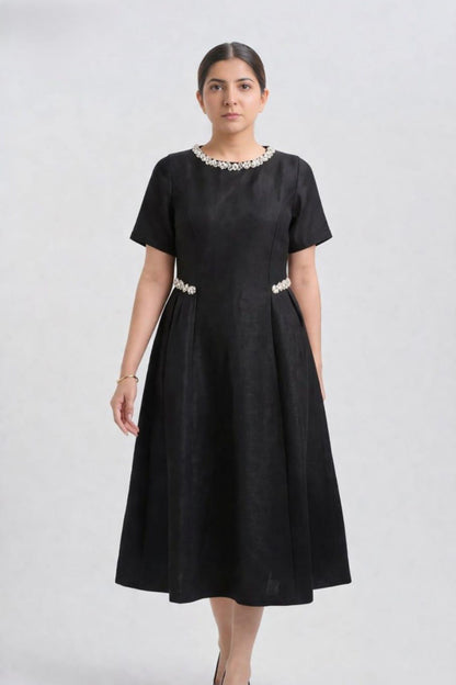 A woman modeling a black short-sleeve midi dress with a beaded neckline and a beaded waistband, set against a plain light gray studio background.