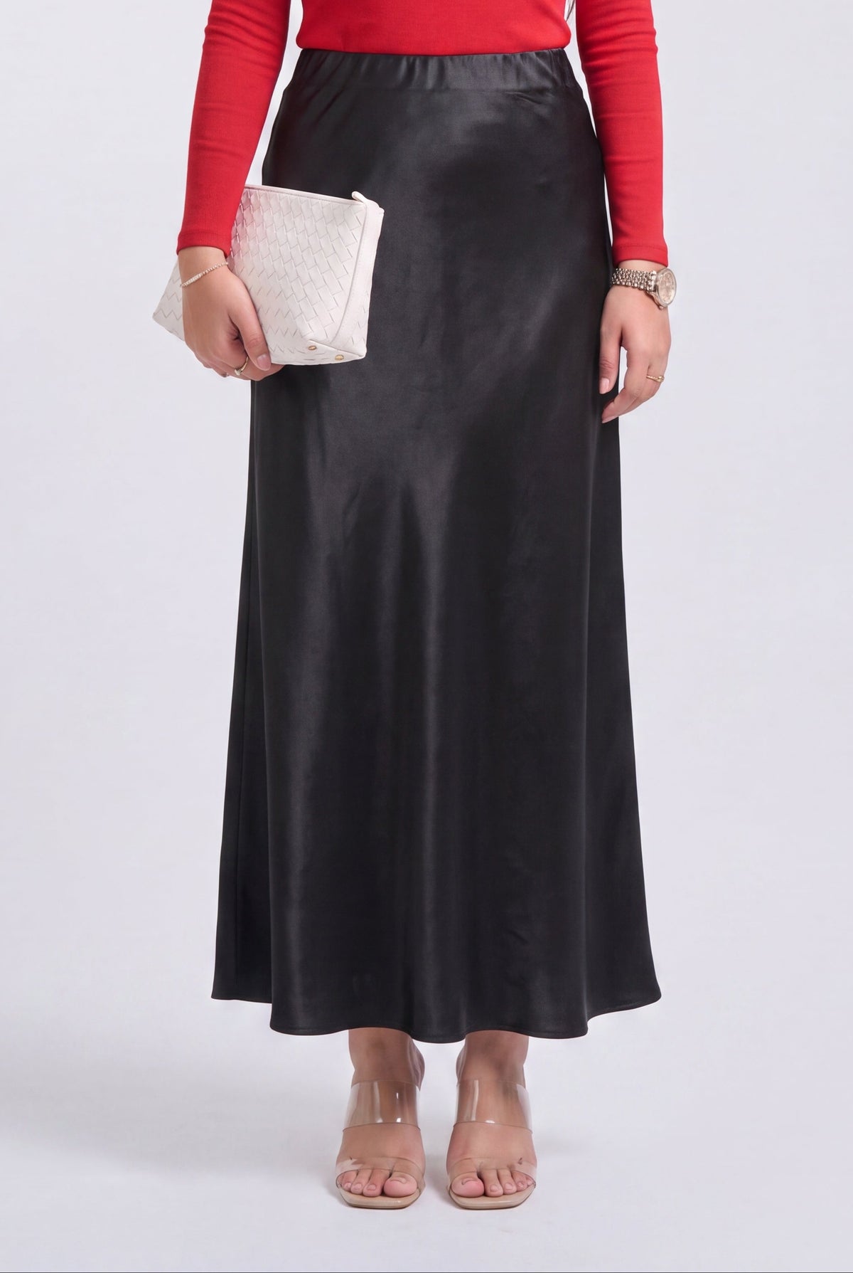 A model wearing a black satin midi skirt, a red long-sleeve top, and clear block-heel sandals, holding a white quilted clutch.