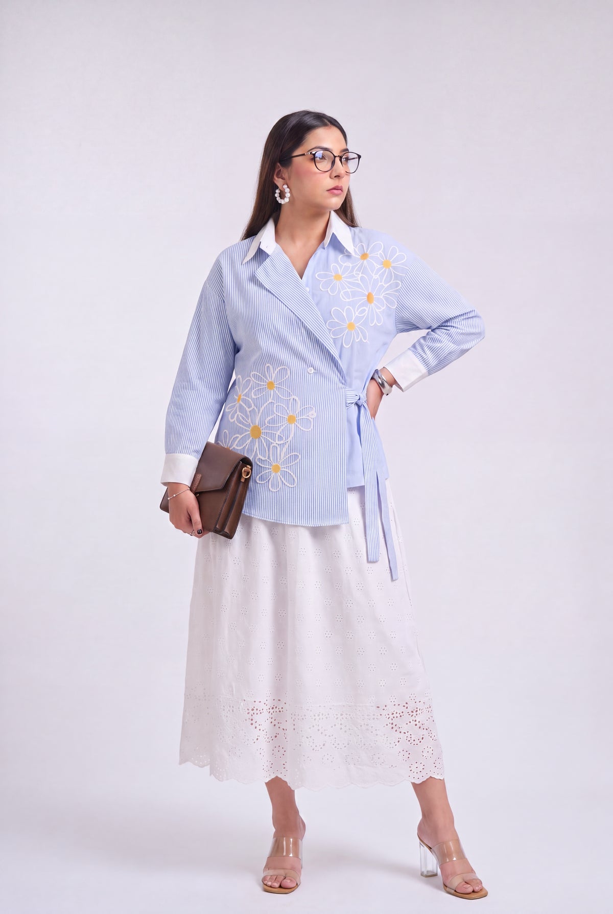 A woman models a light blue long-sleeve embroidered shirt with a white collar and tie waist, paired with a white midi skirt, holding a brown leather clutch against a light gray studio background.