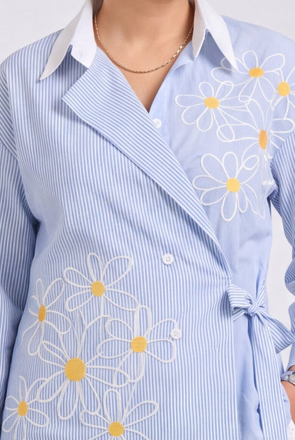 A woman models a light blue wrap-style shirt jacket with white floral embroidery, paired with a white midi skirt and holding a small woven straw bag against a plain light gray studio background.
