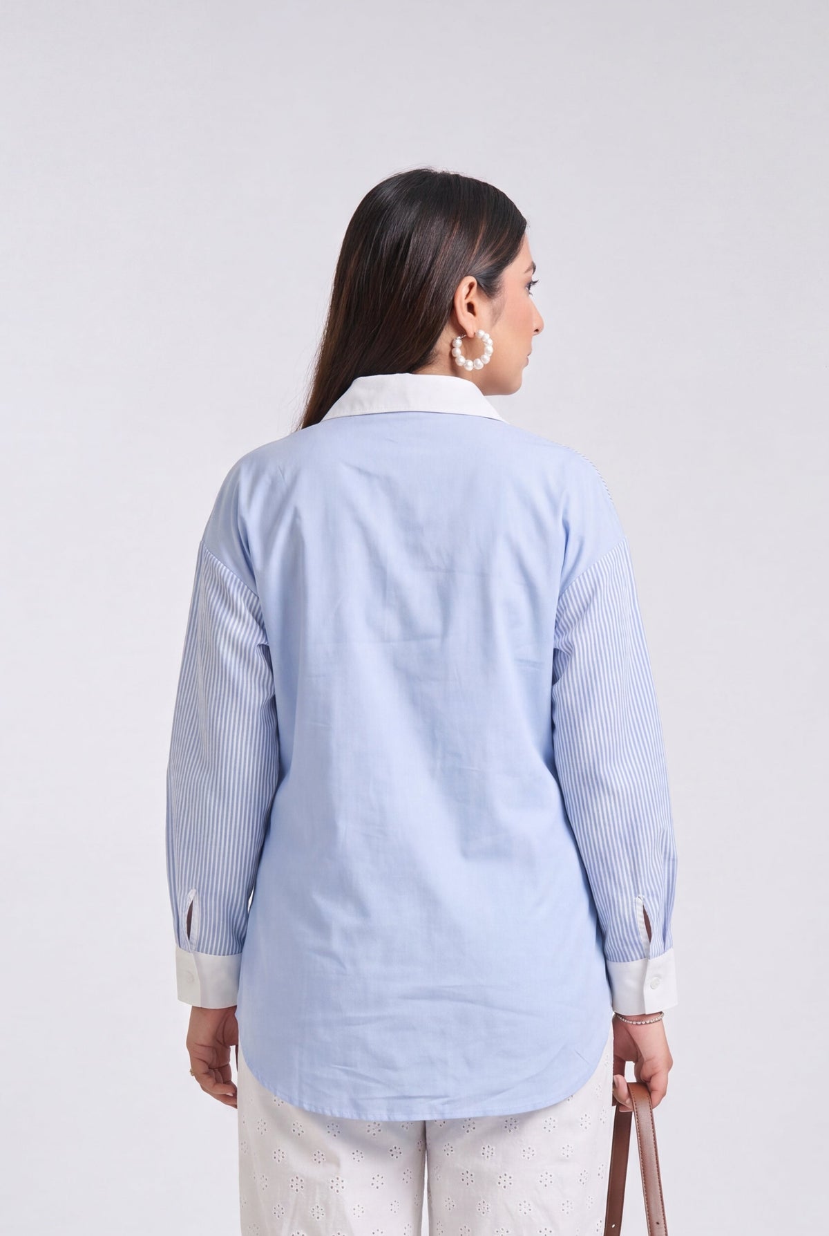 A woman is shown from the back wearing a light blue long-sleeve shirt and white cropped trousers, holding a brown handbag against a plain light gray studio background.