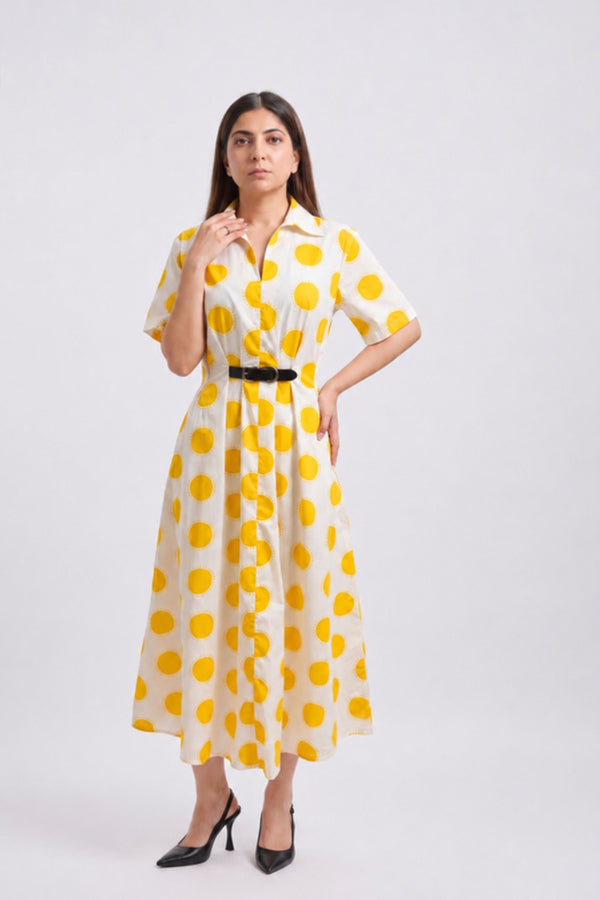 A woman modeling a short-sleeve collared midi dress with a yellow and white polka dot pattern and a black belt, paired with black heels, against a light gray studio background.