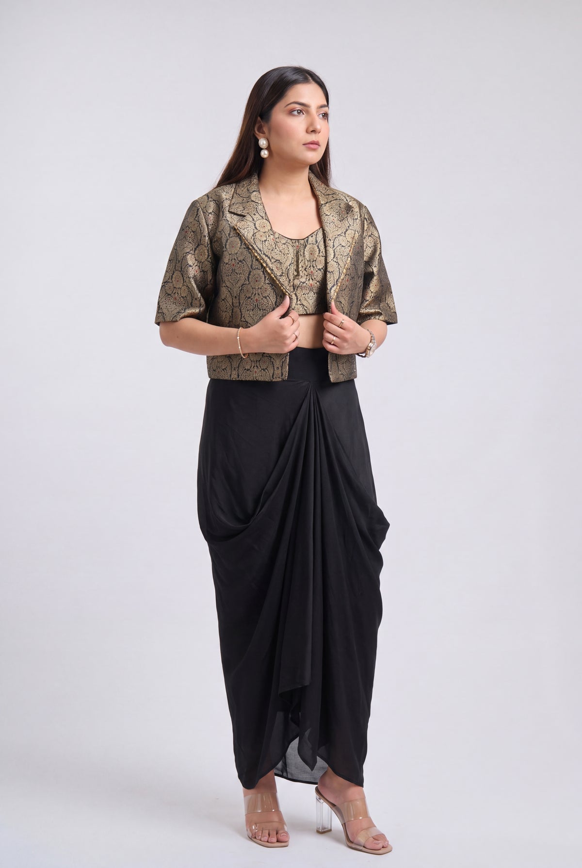 A woman modeling a short-sleeved metallic gold and black brocade jacket paired with a black draped maxi skirt, standing against a light gray studio background.