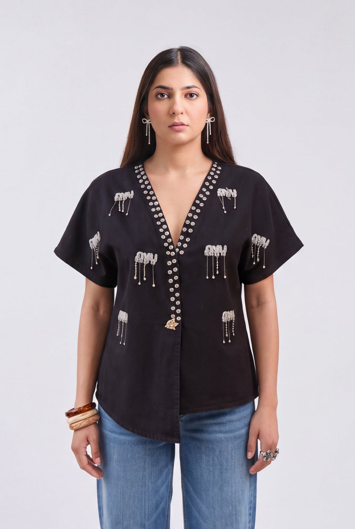 A woman modeling a black short-sleeve button-up blouse with white elephant embroidery and bead trim, paired with blue jeans, against a light gray studio background.