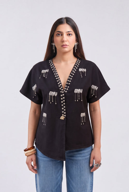 A woman modeling a black short-sleeve button-up blouse with white elephant embroidery and bead trim, paired with blue jeans, against a light gray studio background.