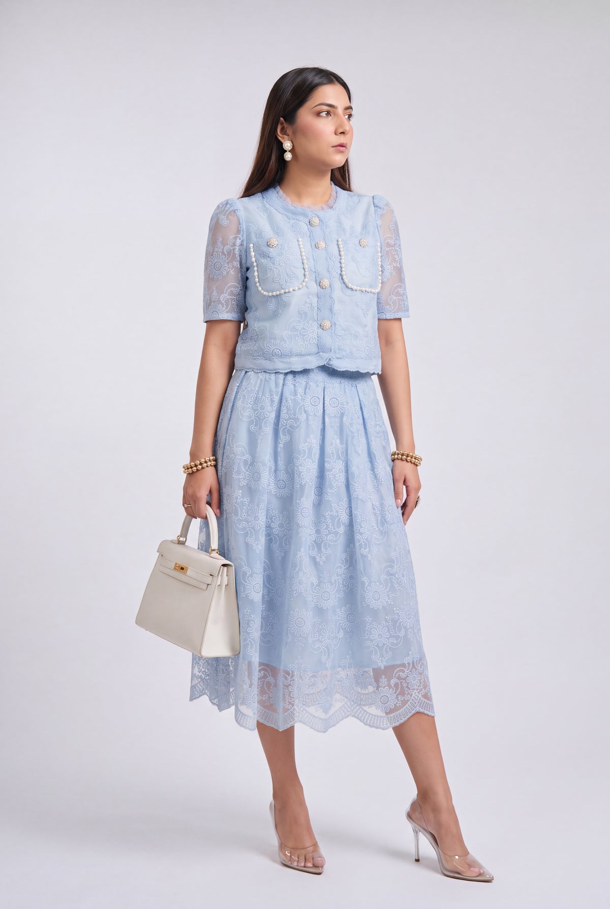 A woman models a light blue two-piece lace set consisting of a short-sleeve cropped jacket with decorative buttons and a matching midi skirt. She is holding a cream-colored handbag and wearing clear high-heeled sandals against a light gray studio background.