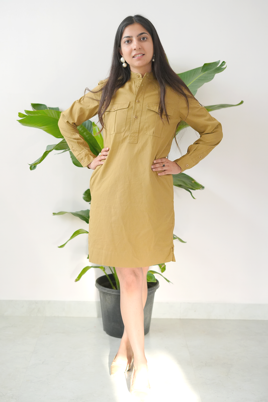 Woman wearing a mustard yellow dress standing in front of a plant against a white wall.