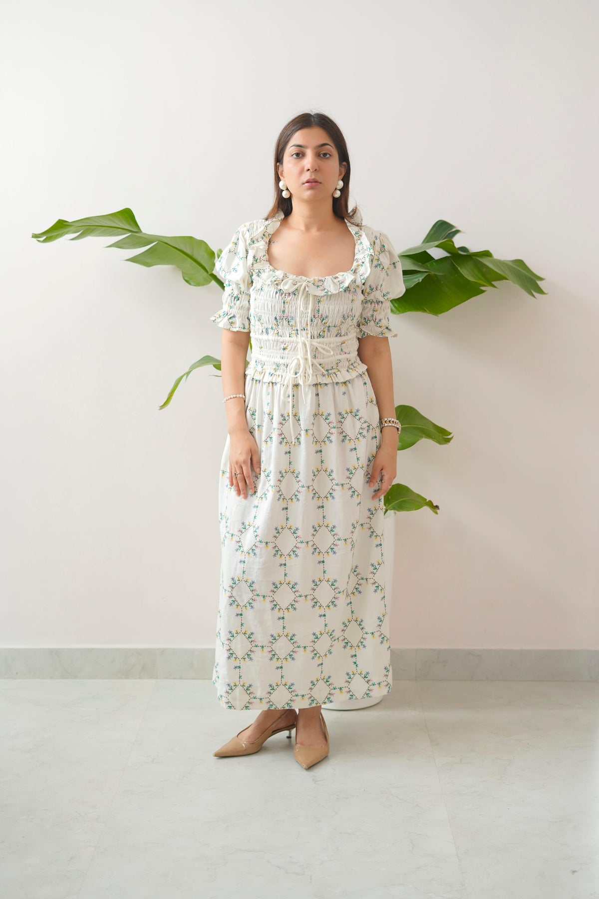 Woman wearing a floral dress standing against a white wall with green leaves.