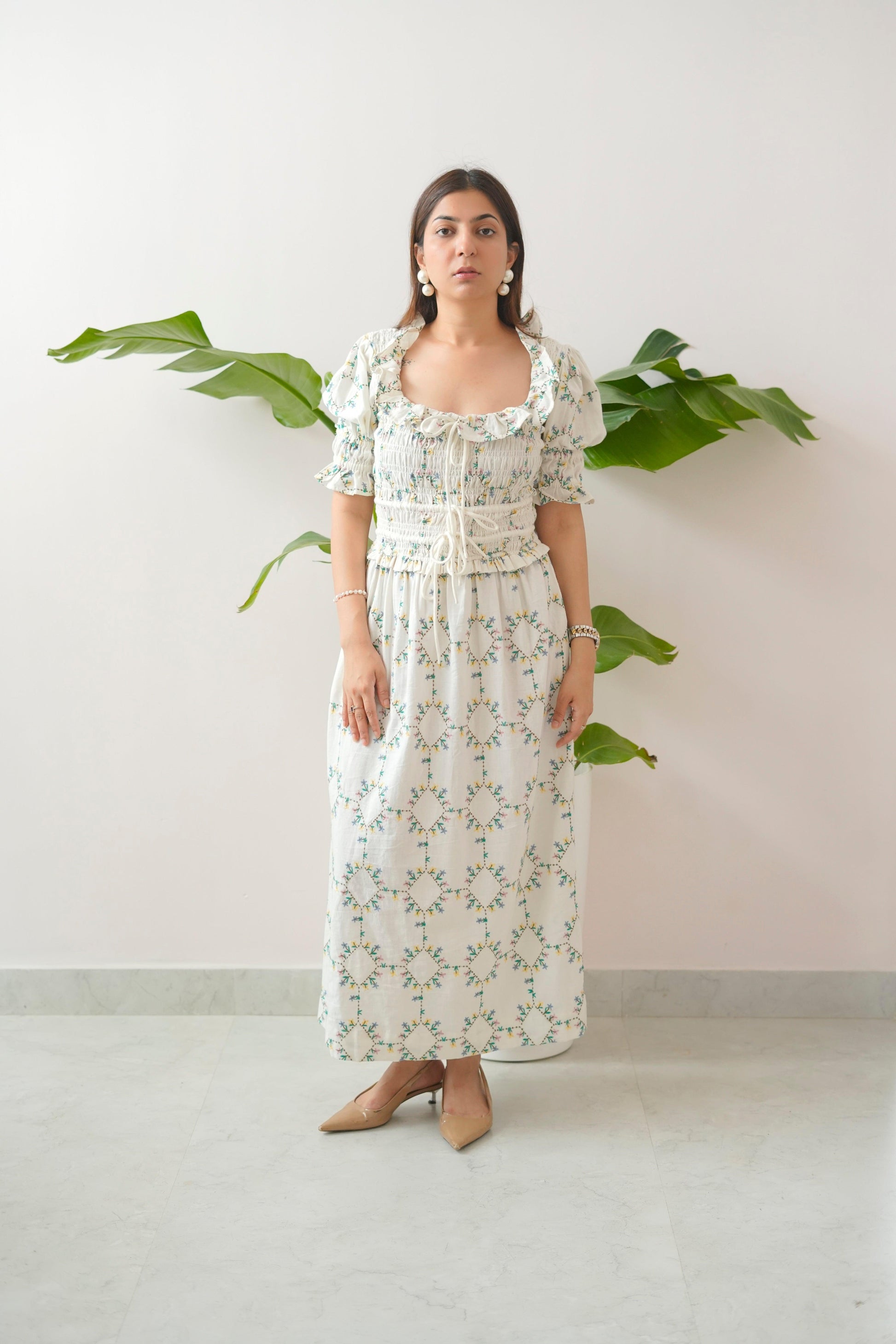 Woman wearing a floral dress standing against a white wall with green leaves.