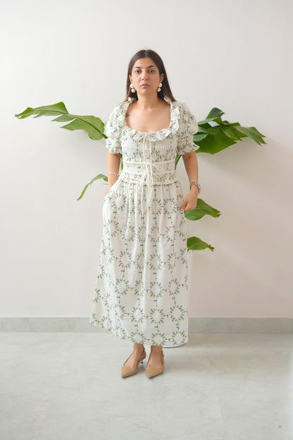 Woman wearing a floral dress standing against a white wall with green leaves.