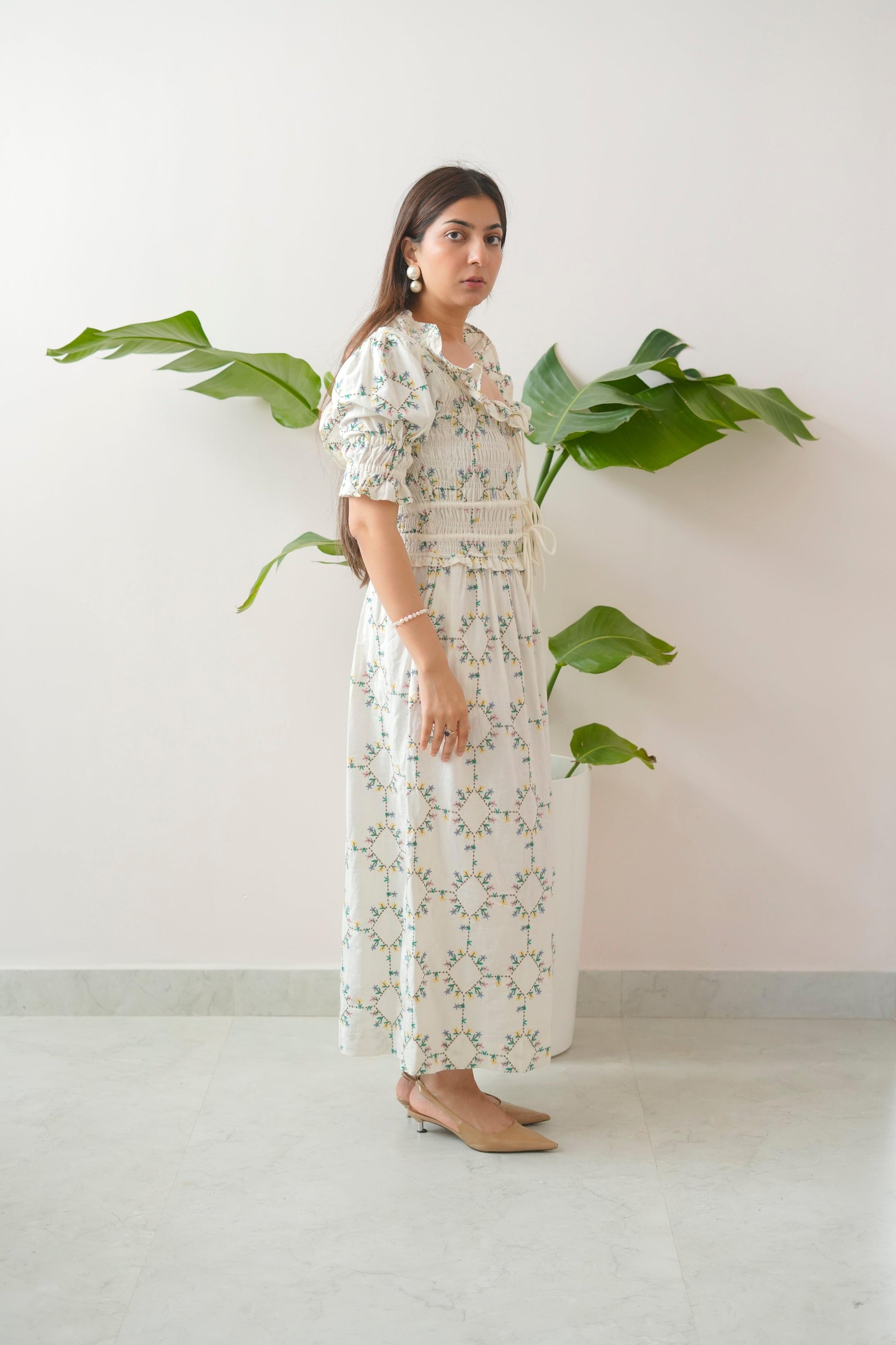 Woman wearing a floral dress standing against a white wall with green plants.