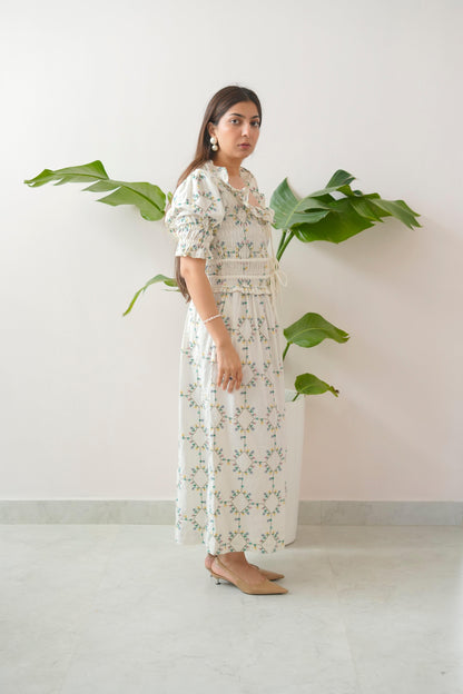 Woman wearing a floral dress standing against a white wall with green plants.