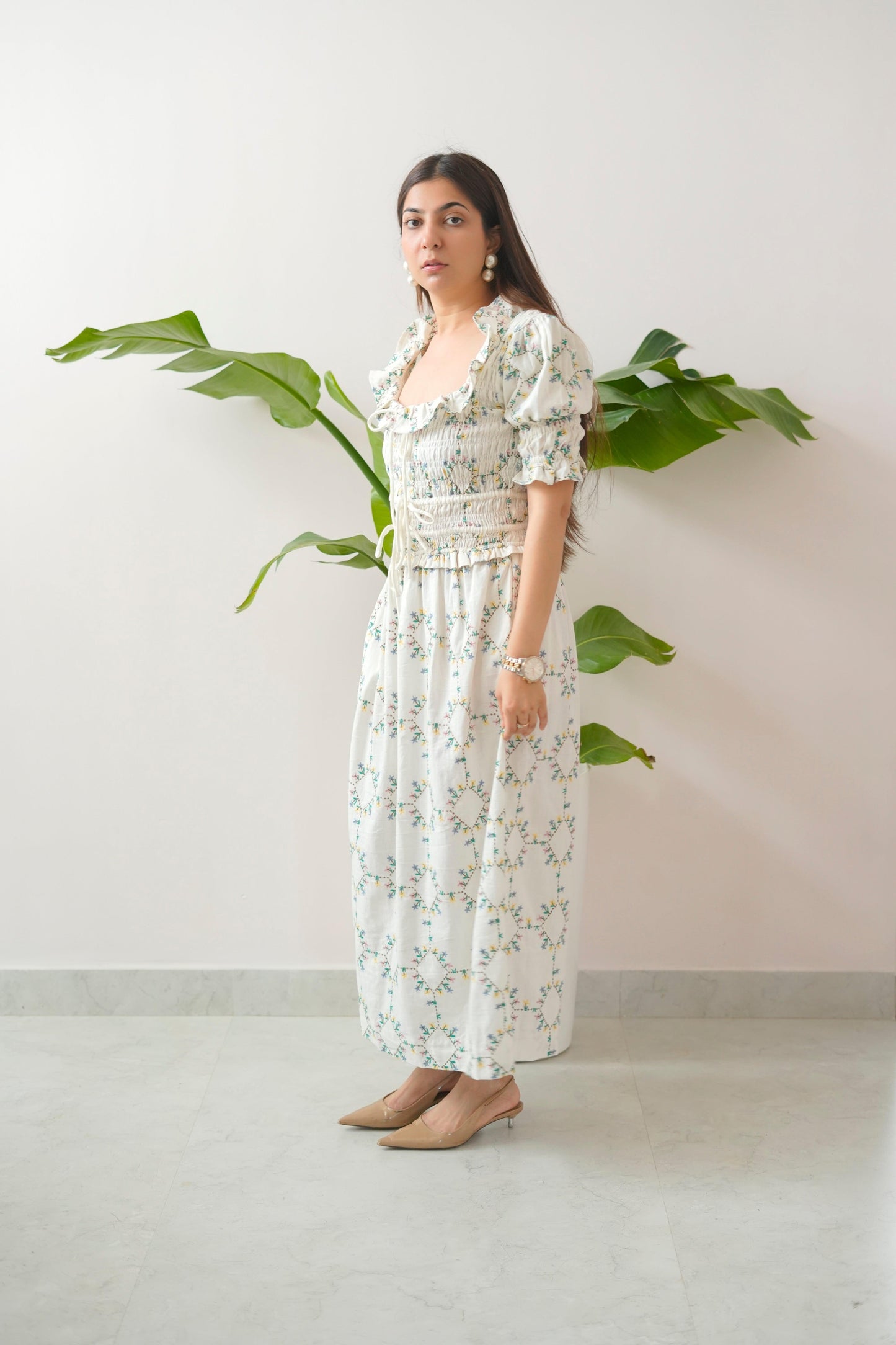 Woman wearing a floral dress standing against a white wall with a plant.