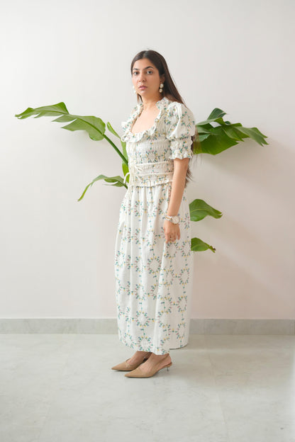 Woman wearing a floral dress standing against a white wall with a plant.