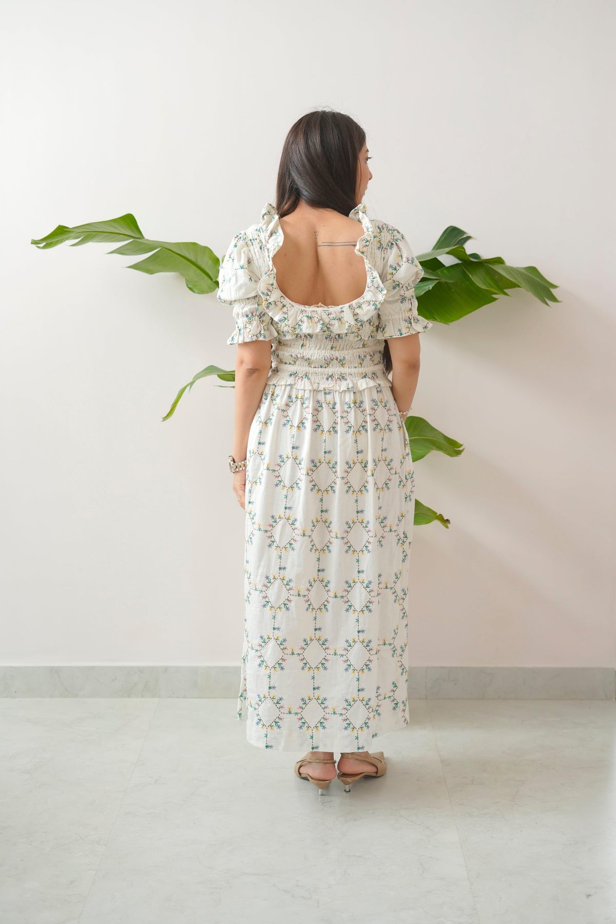 Woman wearing a floral dress standing against a white wall with green leaves.