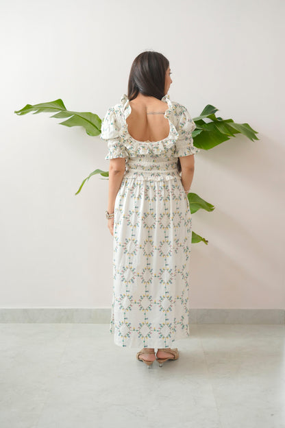 Woman wearing a floral dress standing against a white wall with green leaves.