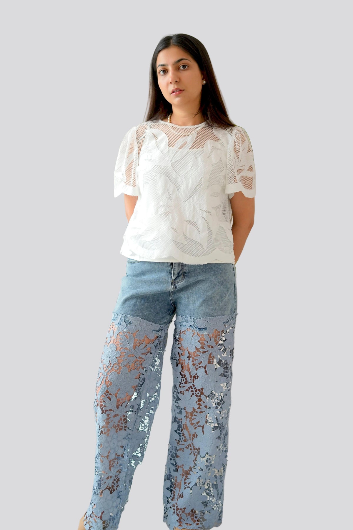 Person wearing a white lace top and blue jeans with lace detailing on a plain background