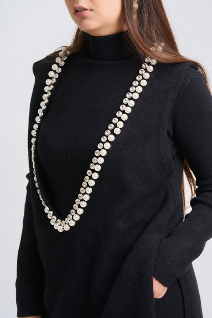 Person wearing a black outfit with a long pearl necklace on a plain background