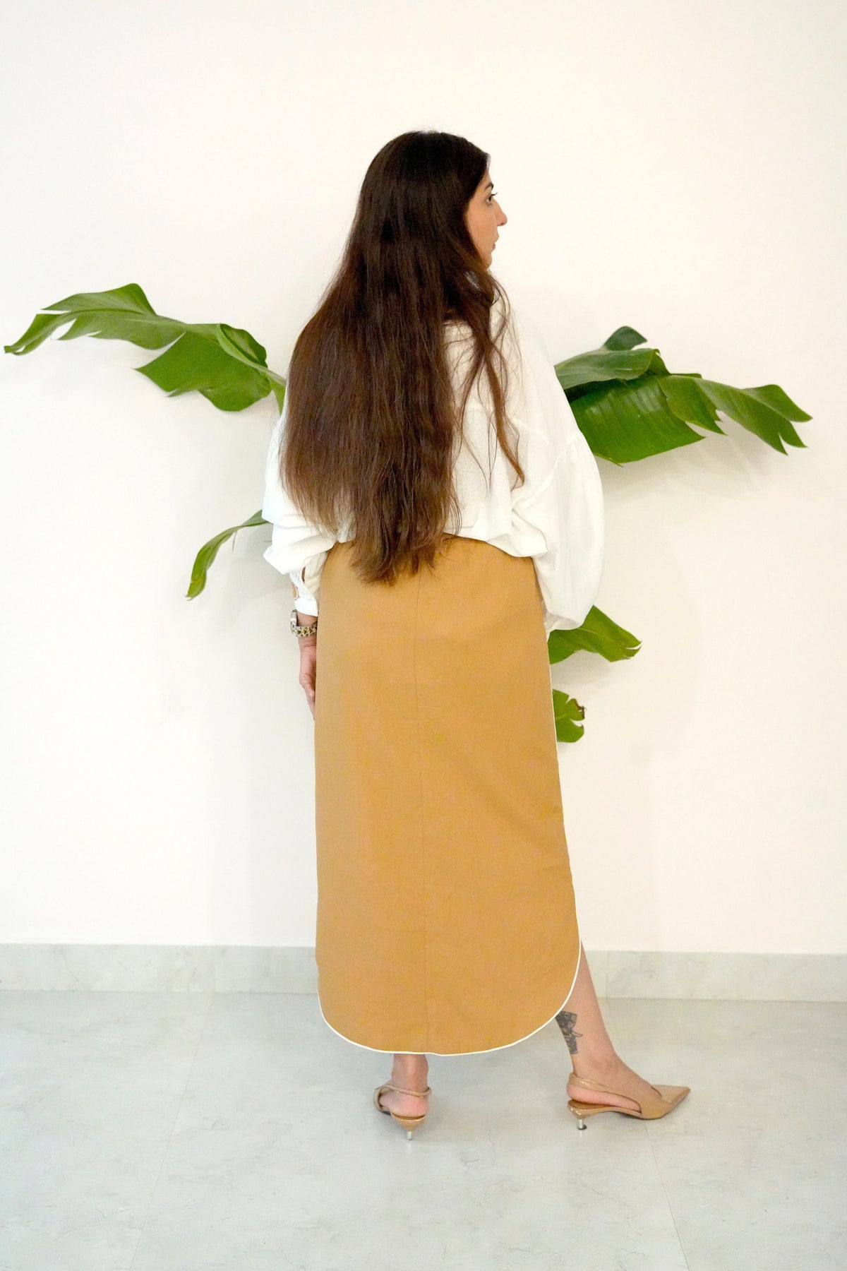 Woman wearing a white blouse and beige skirt standing in front of green plants against a white wall.