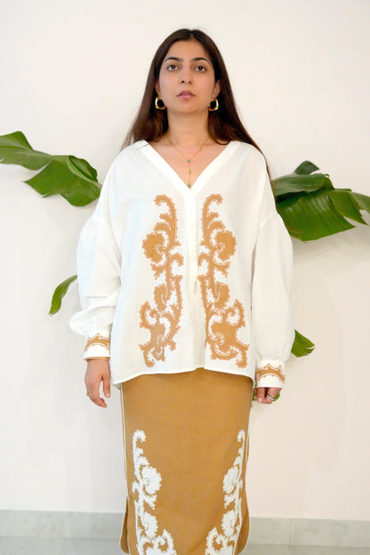 Woman wearing a white blouse with gold embroidery and a brown skirt with white embroidery against a white background with green leaves.