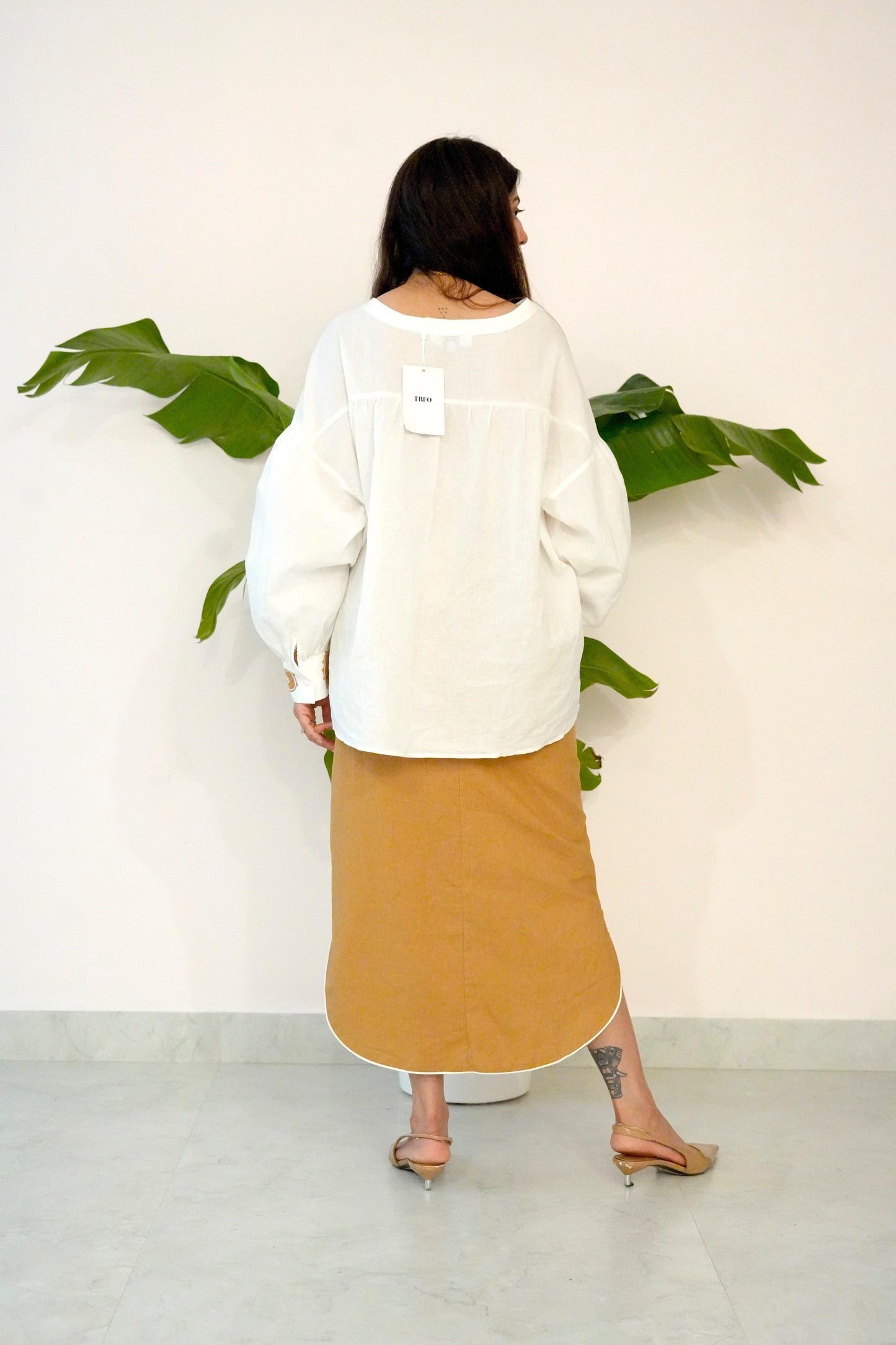 Person wearing a white sweater and beige skirt standing in front of a white wall with green leaves.