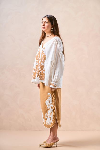 Woman wearing a white blouse with gold embroidery and beige pants against a beige background