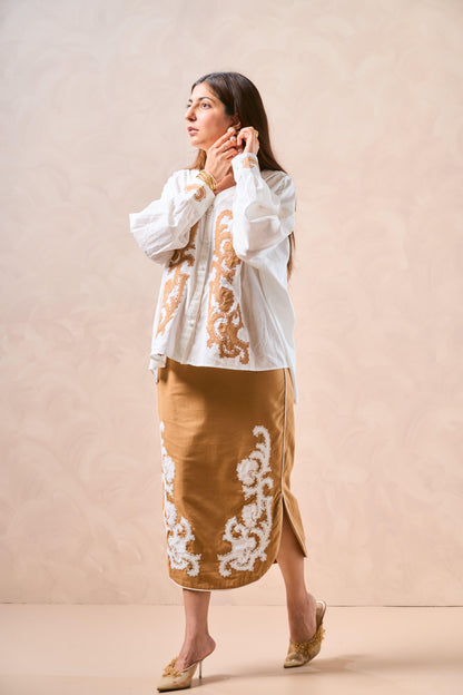 Woman wearing a white blouse with gold embroidery and a brown skirt with white embroidery against a beige background