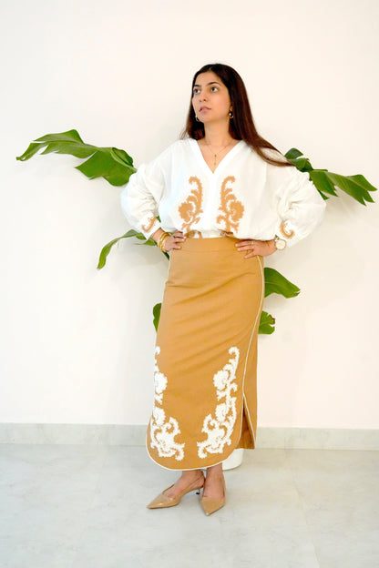 Woman wearing a white blouse with gold embroidery and a beige skirt with white embroidery, standing against a white background with green leaves.