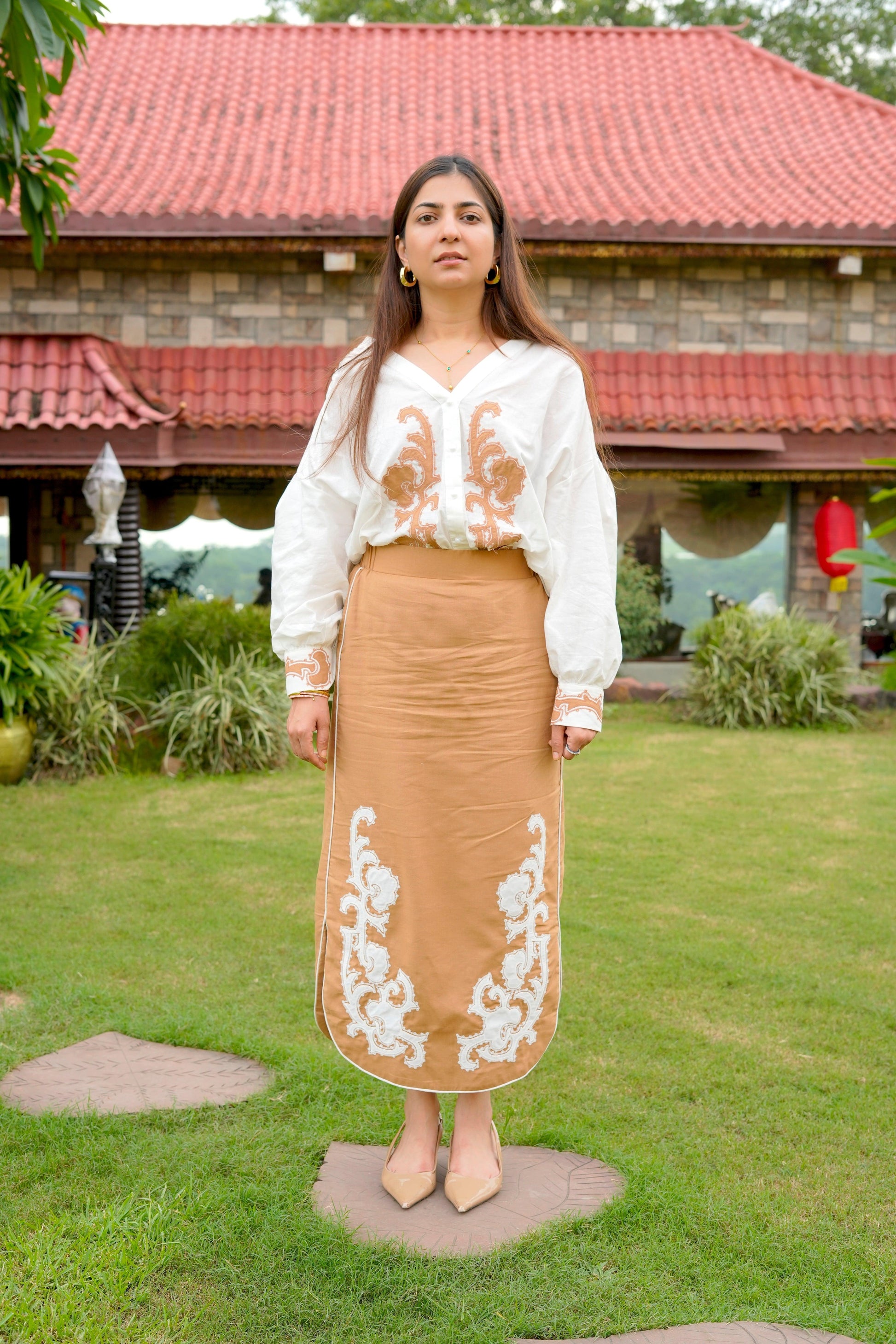 Woman wearing a white blouse with floral embroidery and a brown skirt with white embroidery outdoors.