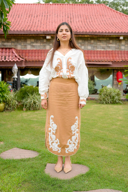 Woman wearing a white blouse with floral embroidery and a brown skirt with white embroidery outdoors.