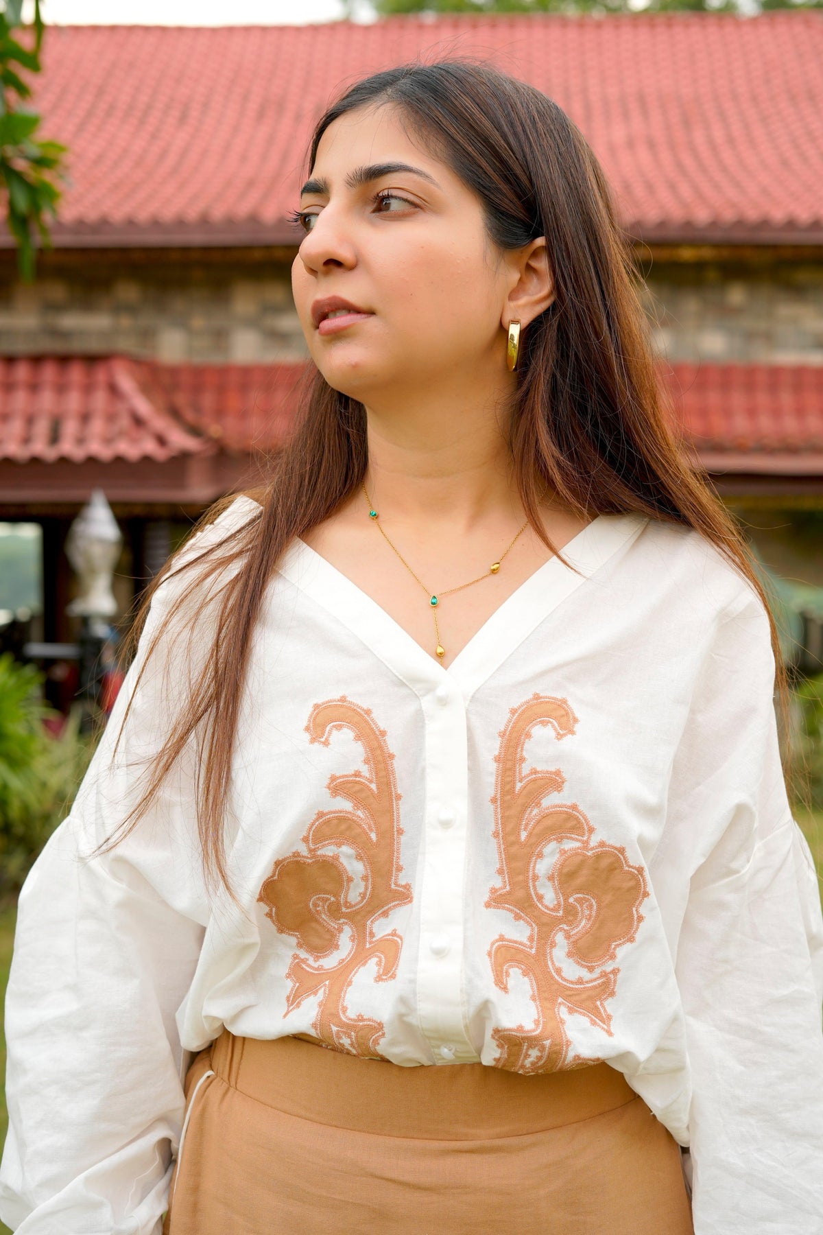 Woman wearing a white blouse with brown embroidery and beige pants outdoors.