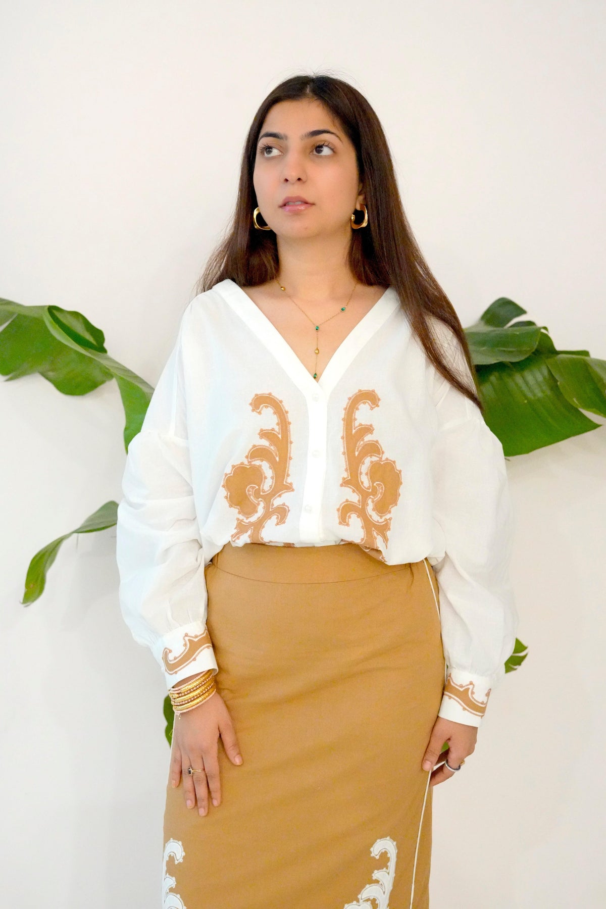Woman wearing a white blouse with gold embroidery and a beige skirt against a white background with green leaves.