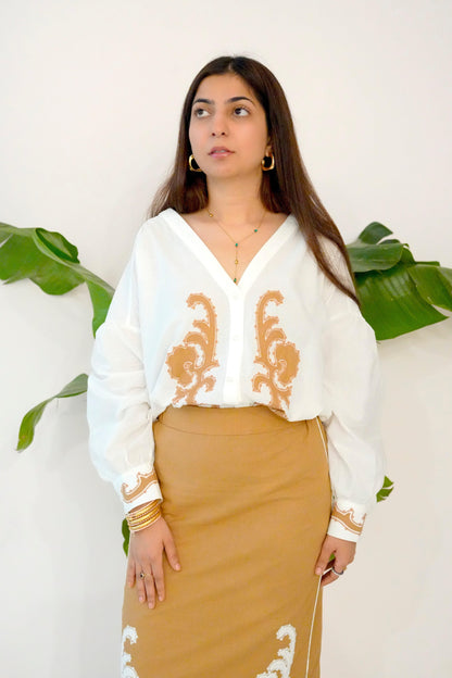 Woman wearing a white blouse with gold embroidery and a beige skirt against a white background with green leaves.