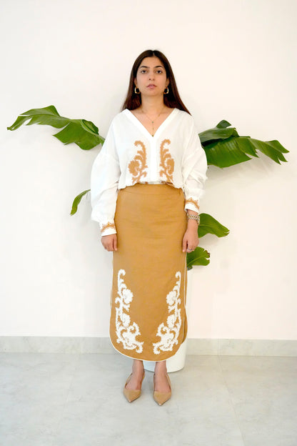 Woman wearing a white blouse and tan skirt with embroidery, standing against a white wall with green plants.